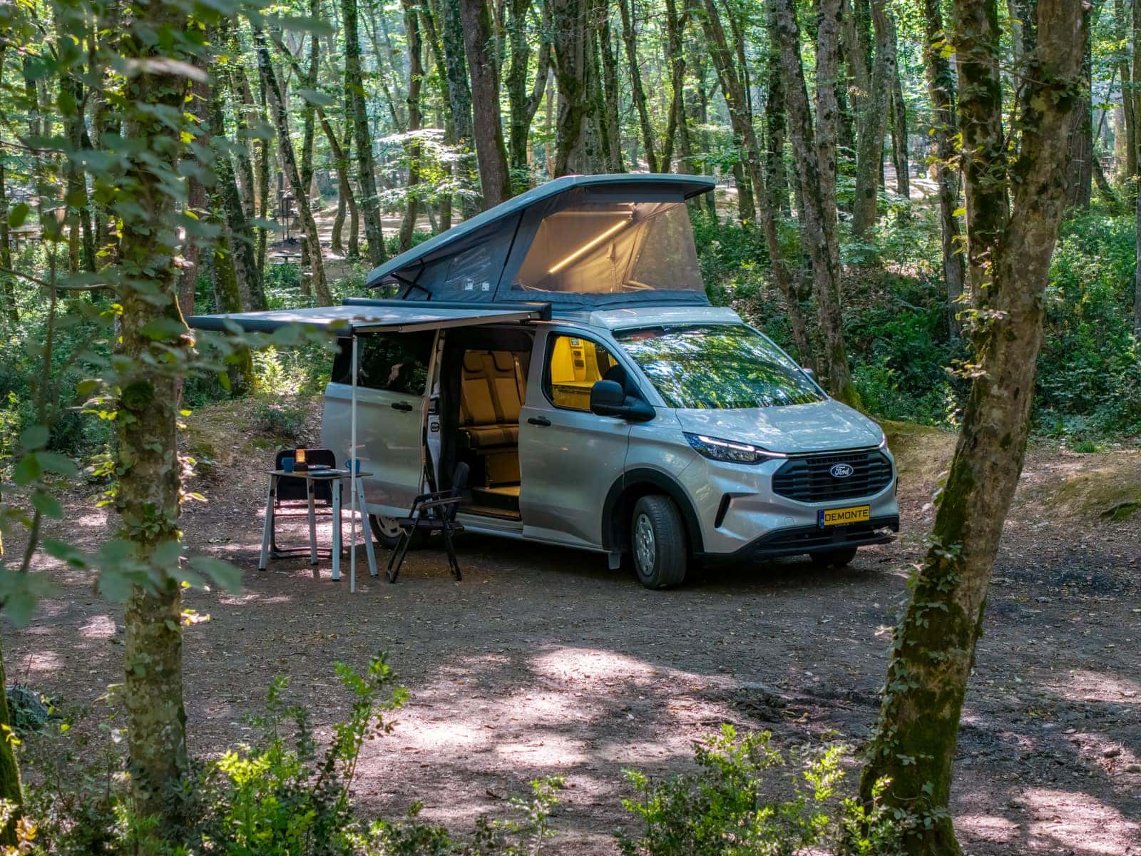 Assos auf Ford Transit Custom im Wald, Aufstelldach und Markise geöffnet, Campingtisch aufgebaut