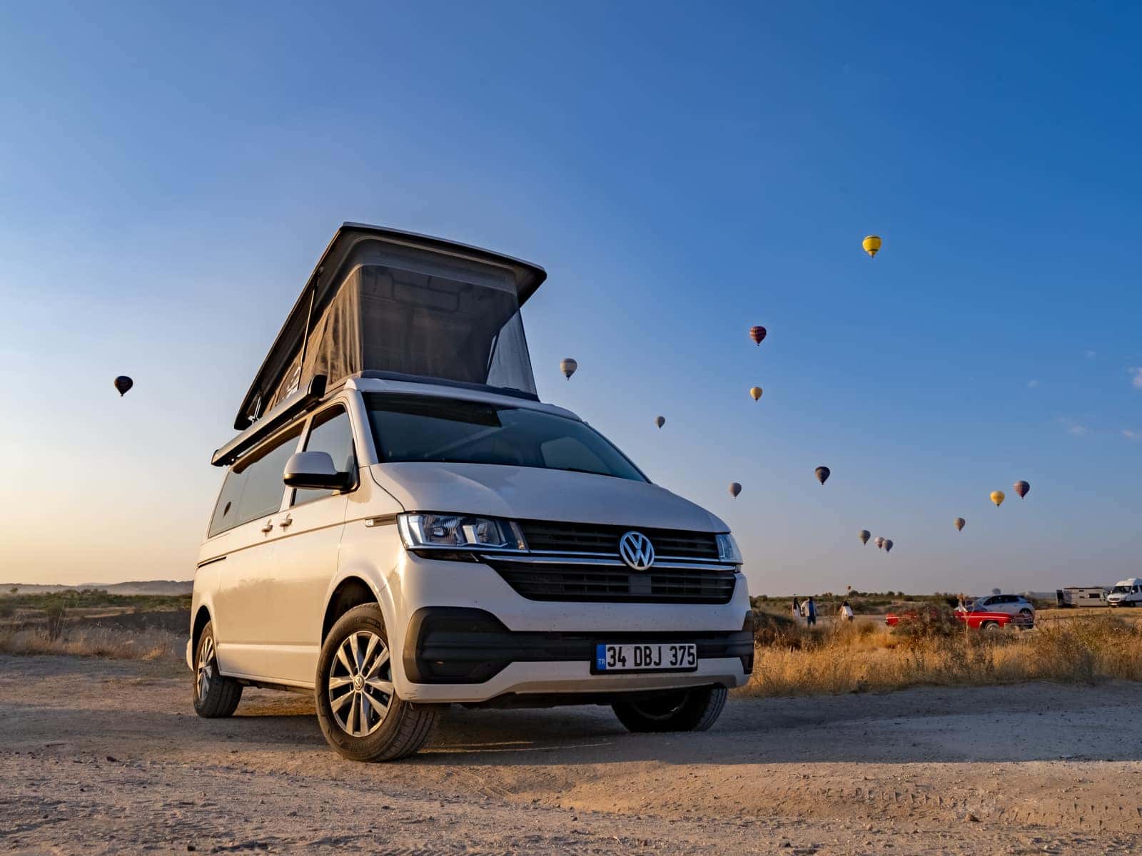 Cappadocia on the white VW T6.1 at sunrise on the Cappadocian plain, pop-up roof open and hot-air balloons in the sky