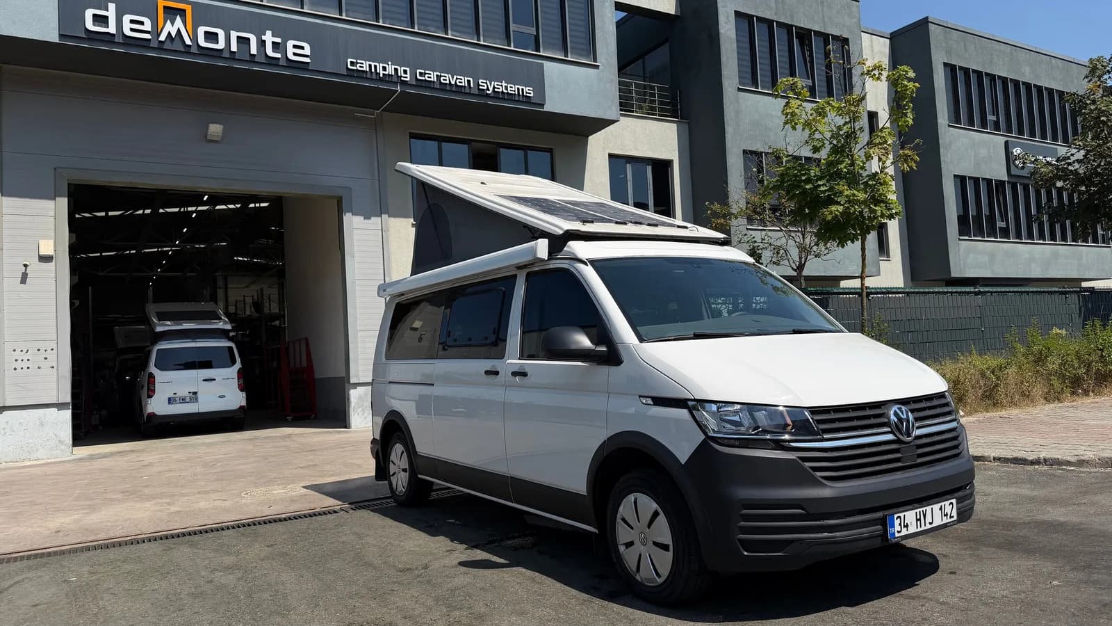 Beyaz VW Transporter Efes — Demonte Caravan üretim tesisi önünde, SCA ters açılır tavan ve güneş paneli