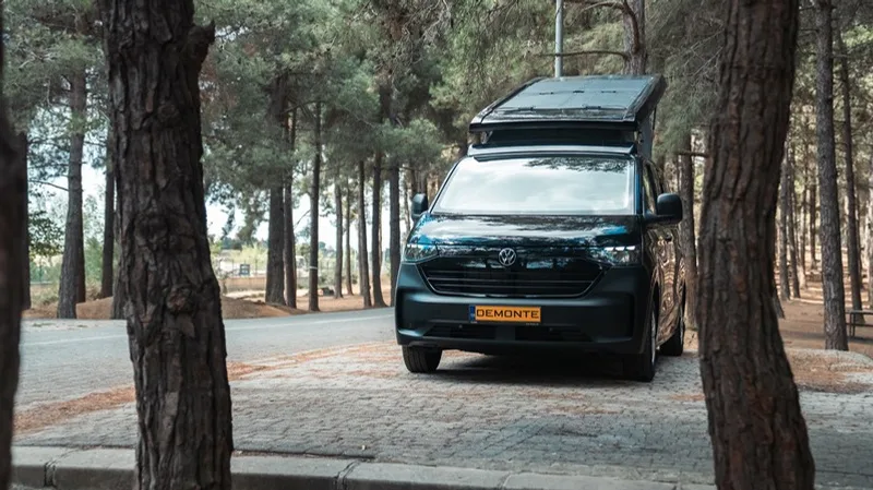 The front of the Olympos framed between two pine trunks, showing the new-generation Transporter grille and the raised roof