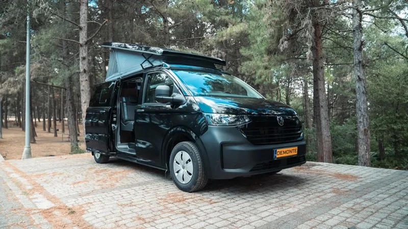 The black Olympos on a paved clearing in a pine forest with the pop-up roof raised and the side door slid open