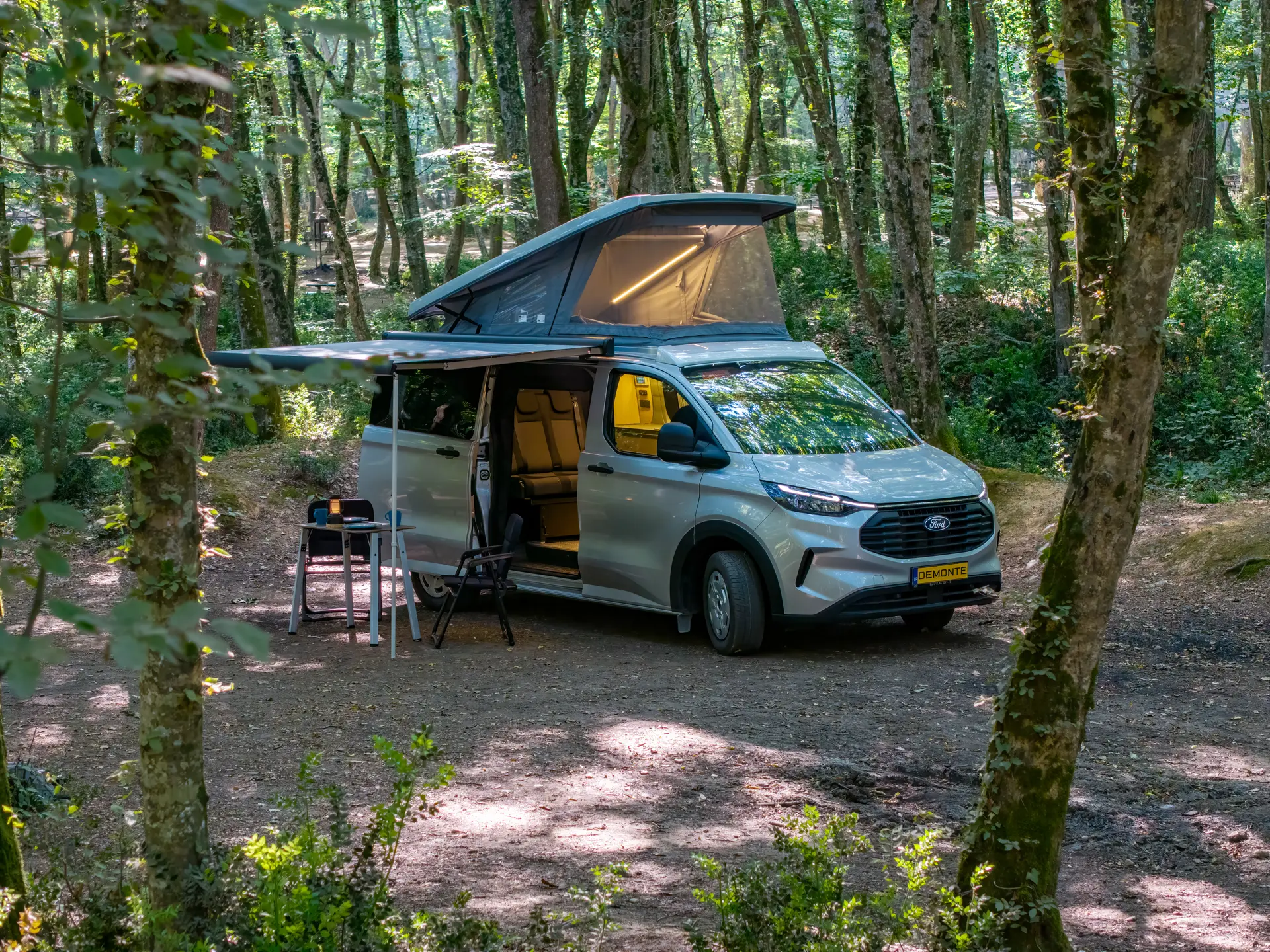 Assos auf Ford Transit Custom, Aufstelldach und Markise im Wald geöffnet, Campingtisch aufgebaut
